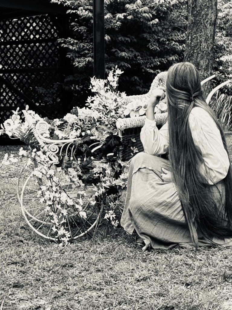A Quiet Moment in the Garden Black and white portrait of a woman in a period dress sitting beside a blossom-covered bicycle in the retreat garden.