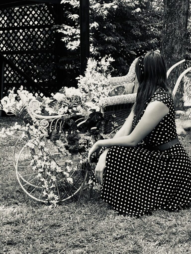 Blossoms and a Bicycle Black and white photo of a woman in a polka dot dress beside a flower-covered garden bicycle and wicker settee.