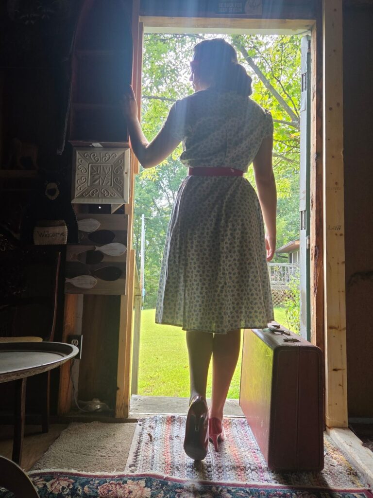 Arriving at the Haven Guest in a vintage dress stepping through the doorway with a suitcase toward the summer lawn at Fisherman's Haven.